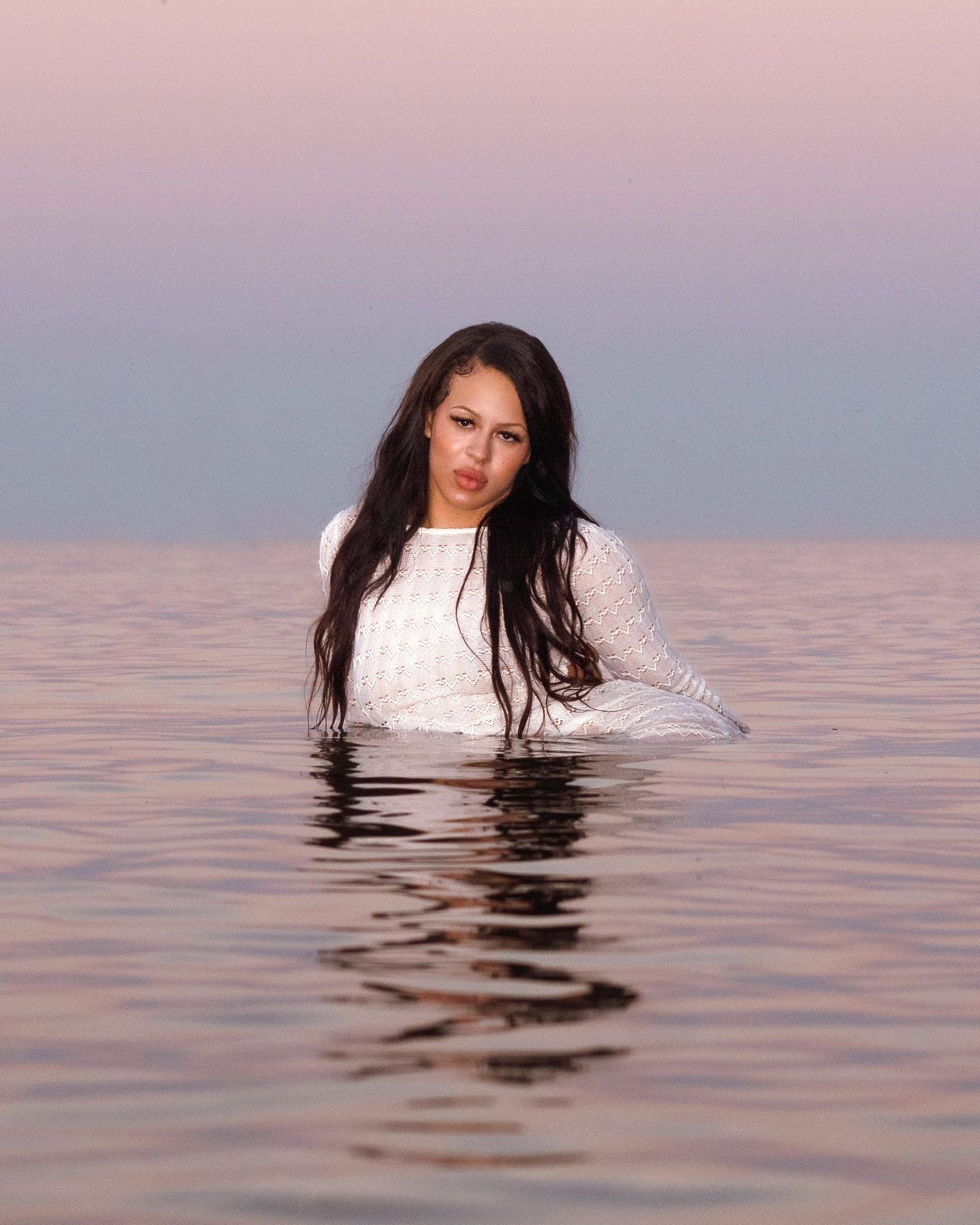 A woman in a white knit top standing waist-deep in still water at dusk, under a pink and grey sky, her reflection broken by ripples.