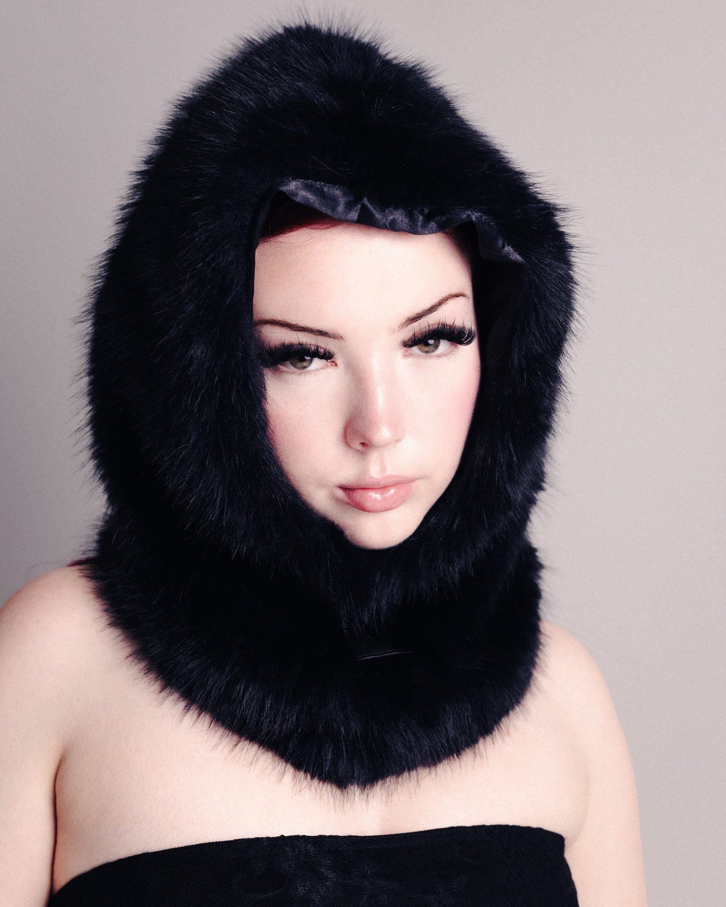 A woman wearing a black fur hood that frames her face, against a pale grey backdrop.
