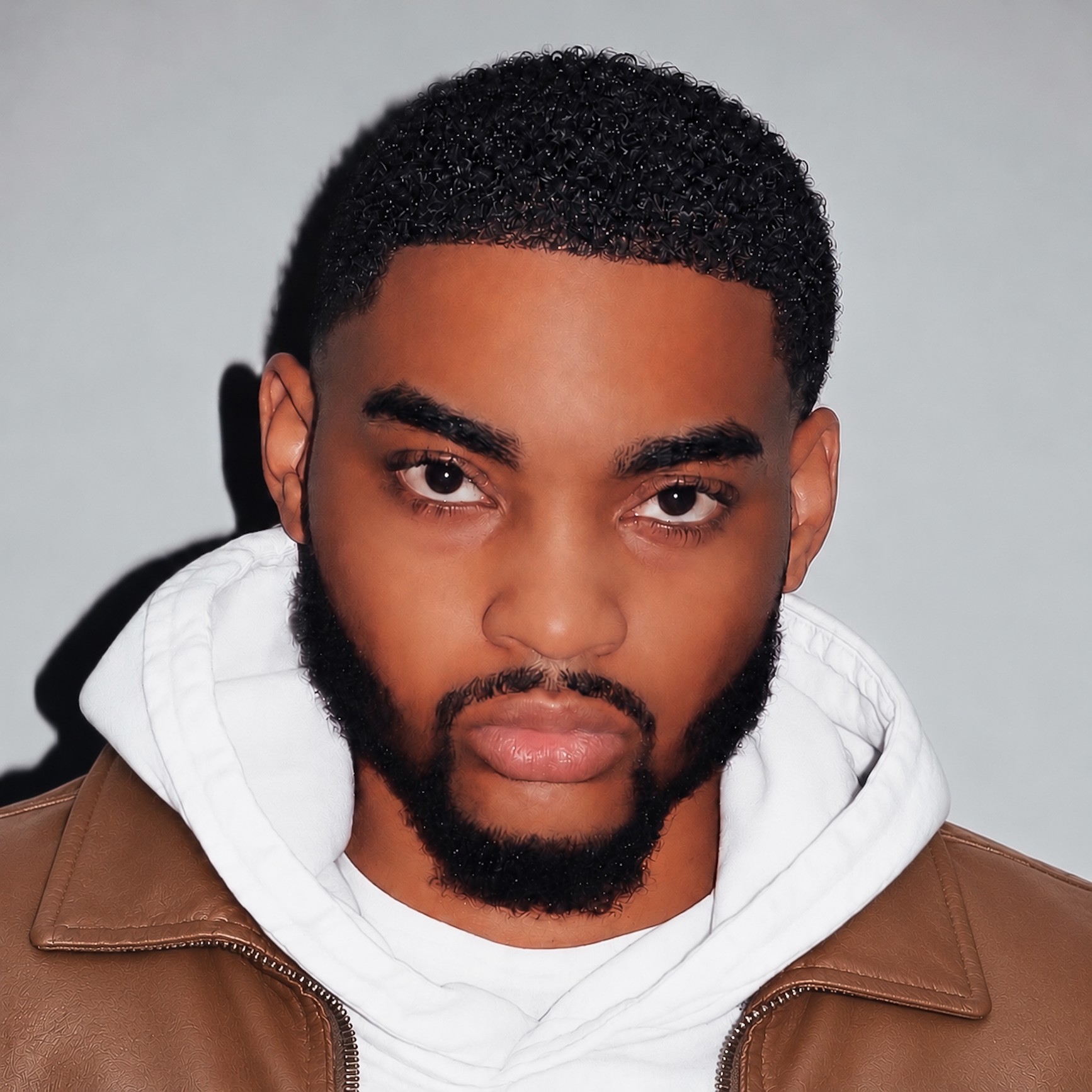 Dontae Kerr, head and shoulders, in a white hoodie and a brown leather jacket against a pale grey wall.