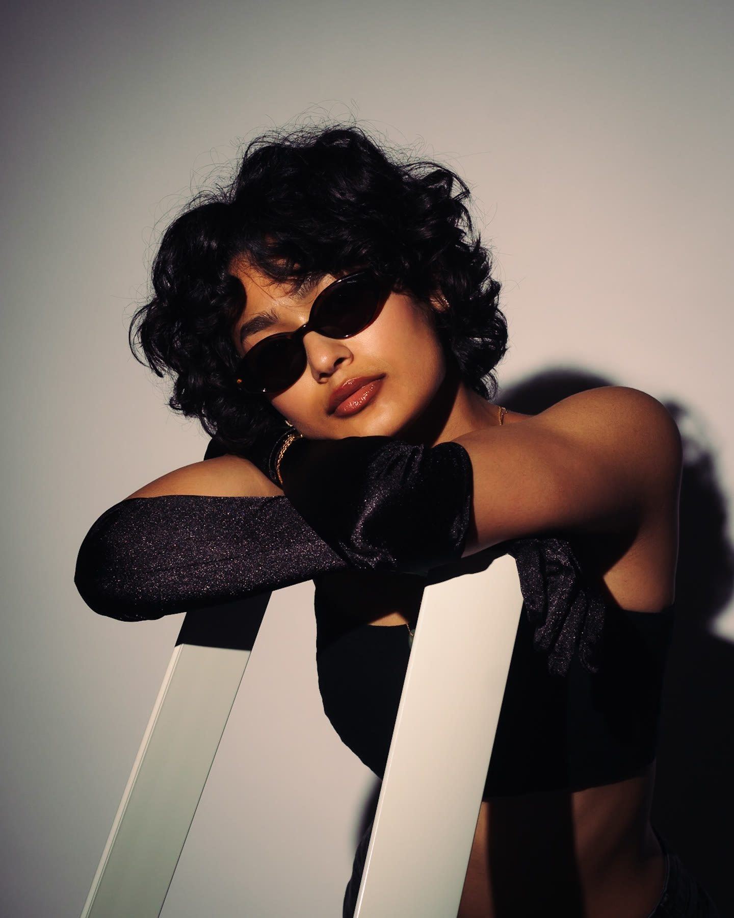 A woman in dark sunglasses and long black gloves leaning over the back of a white chair, her shadow cast on the wall behind her.