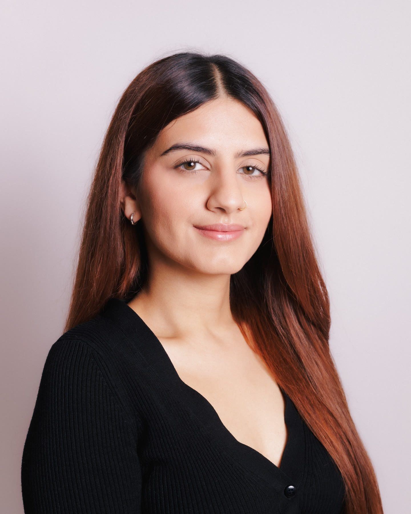 Headshot of a woman with long auburn hair in a black top, with a slight smile, against a soft pink backdrop.