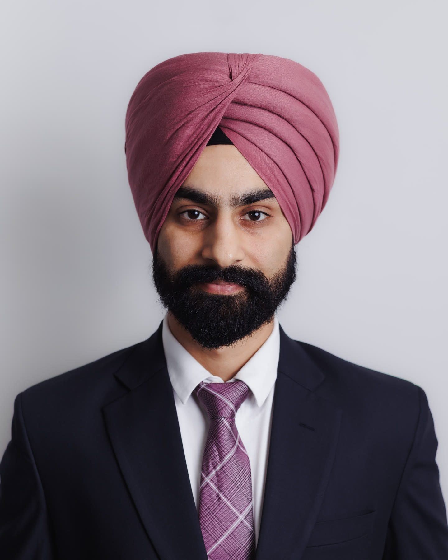 Business portrait of a bearded man in a mauve turban, navy suit and lilac tie, against a pale grey backdrop.