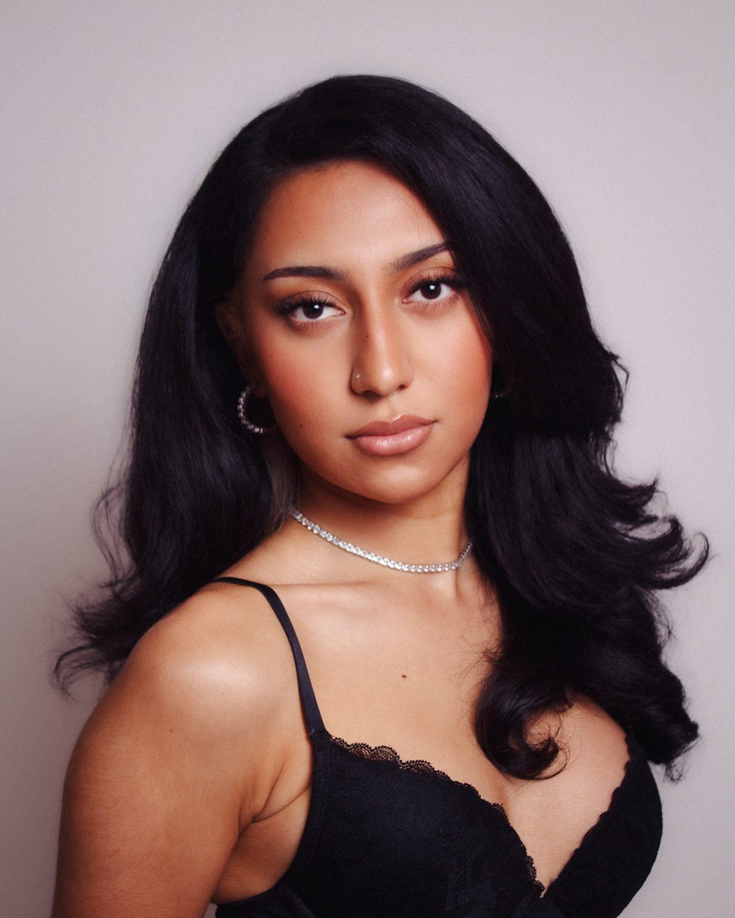 Studio portrait of a woman with long dark hair and a necklace, looking at the camera against a soft pink backdrop.