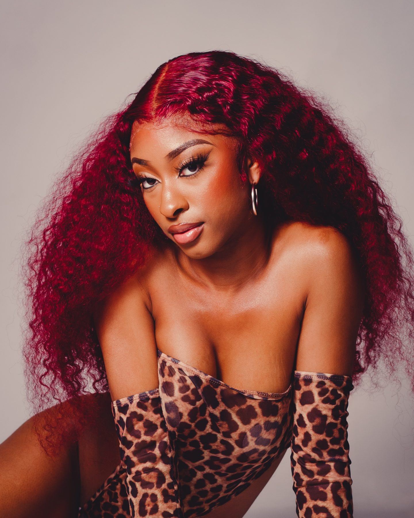 A woman with long crimson curls and leopard-print gloves leaning toward the camera against a grey backdrop.