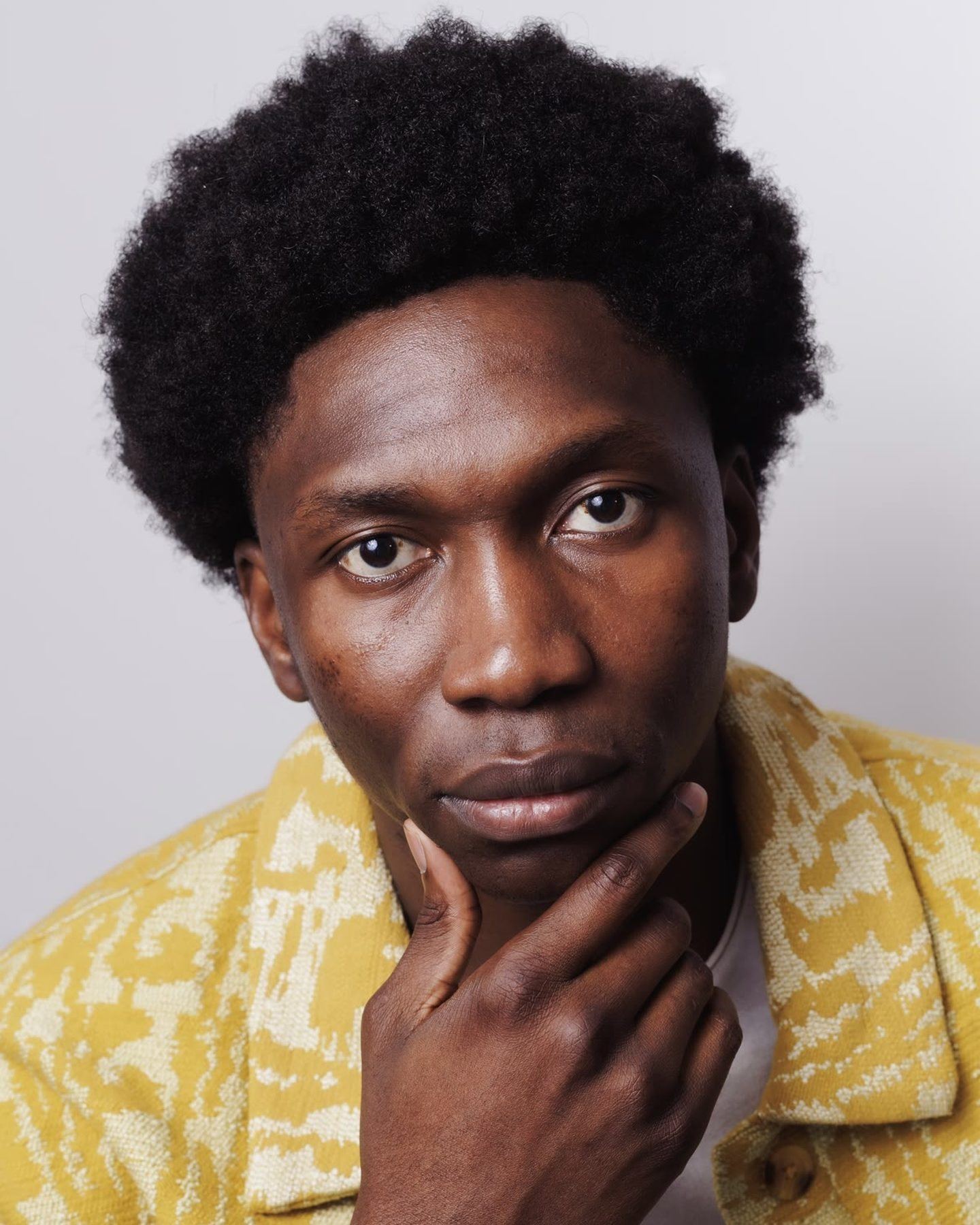 A man with a full afro in a yellow patterned jacket, chin resting on his hand, looking straight at the camera.